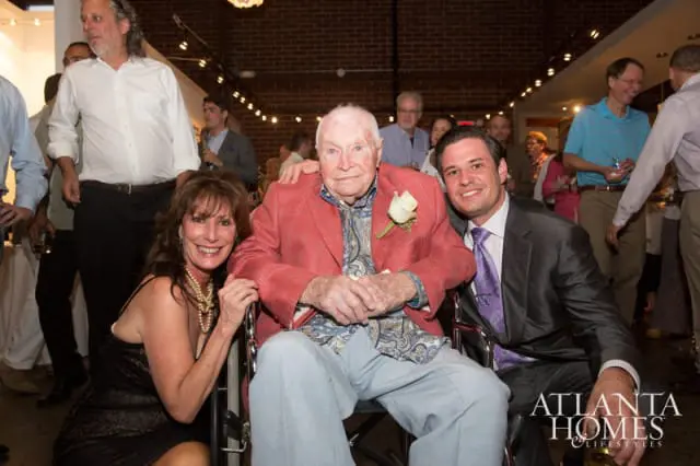 A woman in a black dress and pearls, an older man in a red jacket and patterned shirt in a wheelchair, and a man in a suit and purple tie smile for a photo at an event.