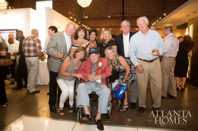 Group of people posing for a photo, with an elderly man in a wheelchair in the center wearing a red jacket and a blue patterned shirt.