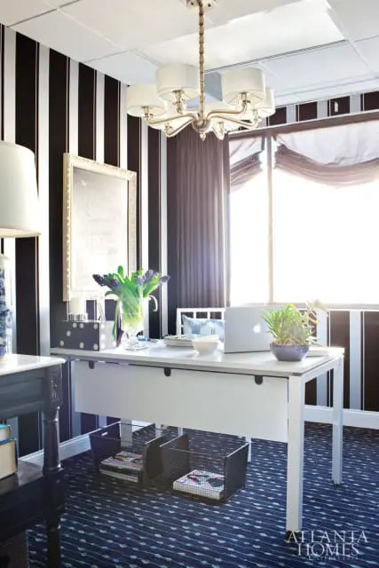 Home office with black and white striped wallpaper, a white desk with a laptop and vase of flowers, and a patterned blue carpet.