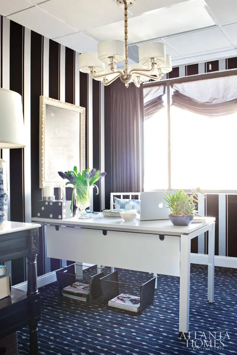 Home office with black and white striped wallpaper, a white desk with a laptop and vase of flowers, and a patterned blue carpet.
