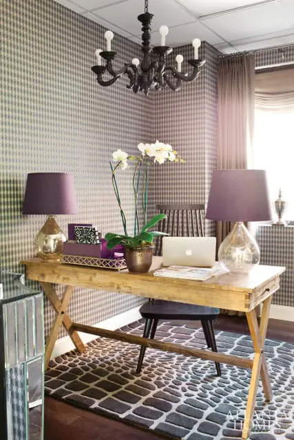 Home office with a wooden desk, purple lamps, a laptop, and a patterned wallpaper. A dark chandelier hangs from the ceiling.