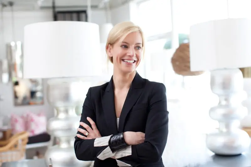 Blonde woman in a black blazer with arms crossed, smiling, with large white lamps and interior decor in the background.