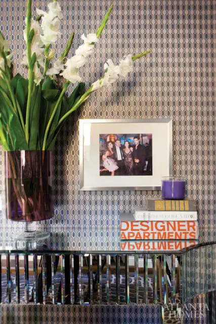 Silver framed photograph of four people in formal attire hangs on a wall with geometric grey and beige wallpaper. A purple vase with white flowers sits to the left, and a stack of books topped with a purple candle sits to the right.