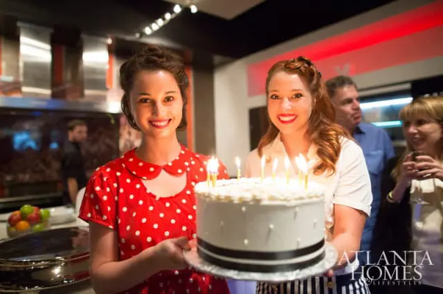 Two women holding a white birthday cake with lit candles. The woman on the left wears a red polka dot dress, and the woman on the right wears a white blouse.