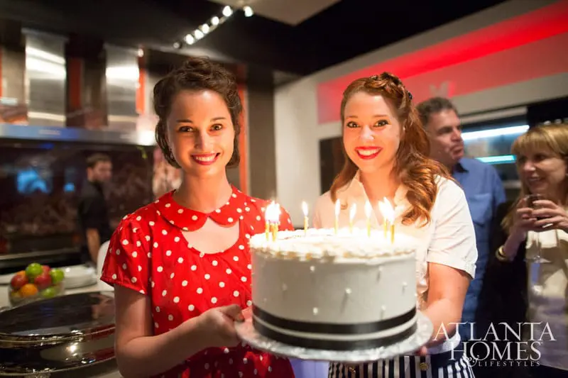 Two women holding a white birthday cake with lit candles. The woman on the left wears a red polka dot dress, and the woman on the right wears a white blouse.