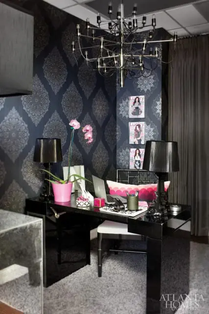 Dark damask wallpaper adorns an office with a black lacquer desk, pink orchid, and a modern chandelier. Three magazine covers are displayed on the wall.