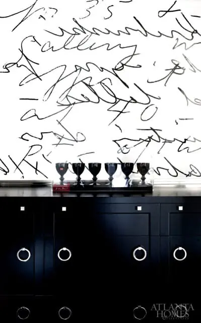 Black dresser with silver ring pulls and square knobs, topped with a black tray holding five black glass goblets and a red book, against a white wall with black abstract calligraphy.