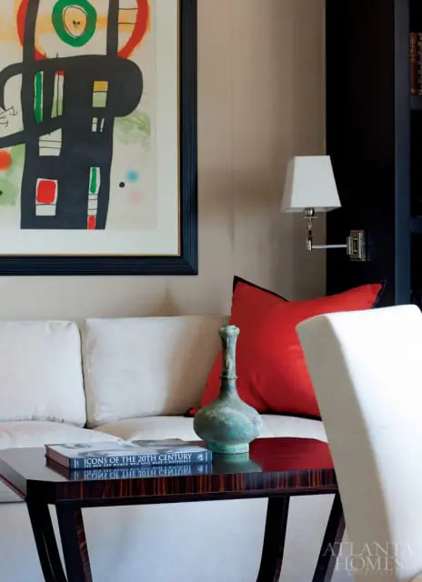 Living room with a beige sofa, red accent pillow, abstract art, and a dark wood table with a book and vase.
