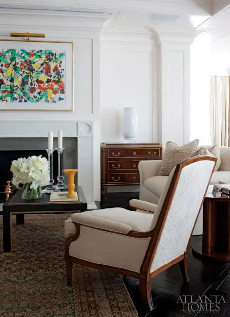Living room with a white fireplace featuring abstract art, a dark wood coffee table with flowers and candles, and upholstered chairs.