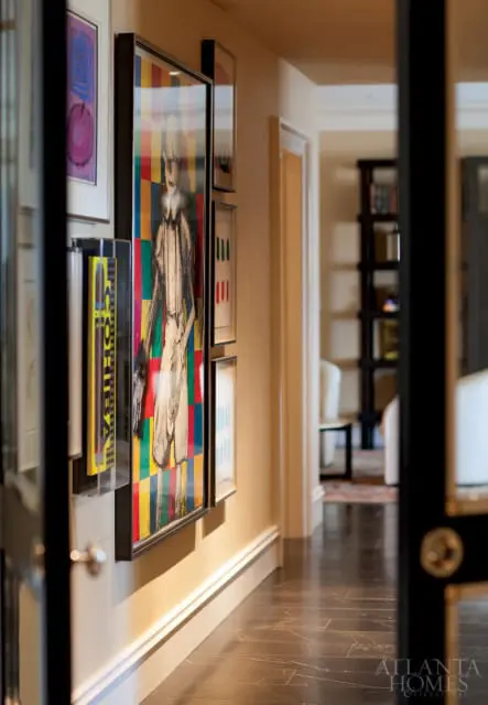 Hallway with framed artwork including a colorful abstract piece and a figure drawing, dark wood floors, and a glimpse of a bookshelf in the background.