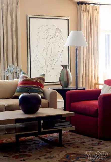 Living room with a tan sofa, red armchair, and a coffee table with a dark apple sculpture. A large framed line drawing hangs on the wall behind a striped vase.