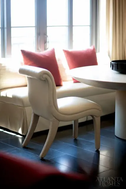 Cream upholstered dining chair with curved back and textured legs at a round white table next to a banquette with coral pillows.