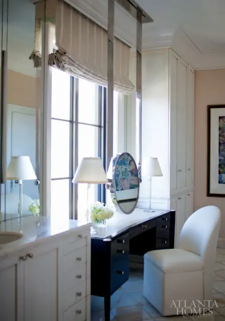 Vanity area with a black lacquer desk, white chair, marble countertop, and mirrored wall panels. A large window with striped roman shades is behind the desk.