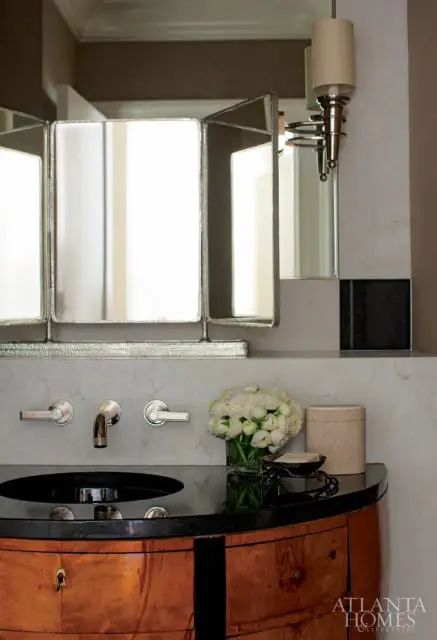 Bathroom vanity with a black granite countertop, wood cabinetry, marble backsplash, and a triptych mirror. A vase of white roses sits on the counter.