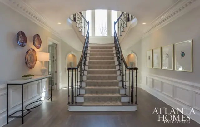 Foyer with a grand staircase featuring a patterned carpet runner and dark wood banisters, flanked by arched niches and artwork. A console table with a lamp and decorative plates adorns the left wall.