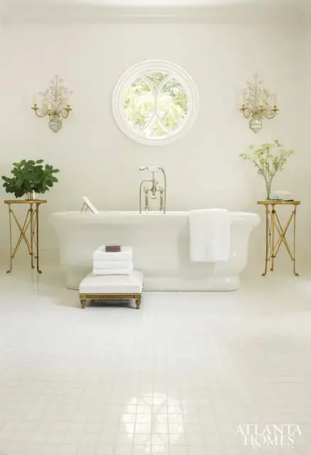 White bathroom with a freestanding tub, round window, mosaic tile floor, and ornate sconces. A stack of white towels sits on a decorative ottoman.