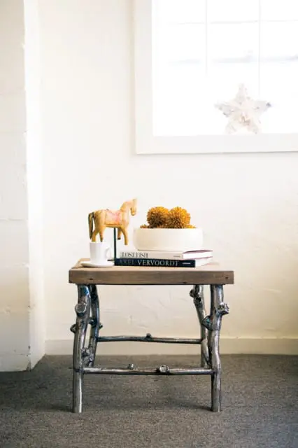 Wooden side table with a rustic metal base, topped with a white mug, a wooden horse figurine, and a white bowl filled with orange decorative spheres.