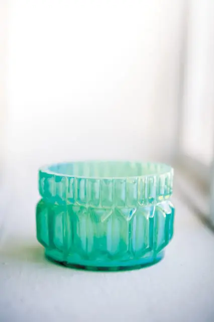 Close-up of a teal iridescent glass votive holder with geometric facets and a ribbed rim, resting on a white surface.