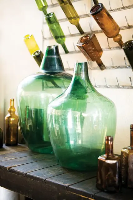 Collection of large green glass demijohns and smaller amber glass bottles displayed on a dark wooden shelf with a rack of bottles in the background