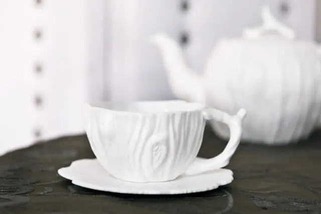 White ceramic teacup and saucer with a textured, wood-grain design, with a matching teapot blurred in the background.