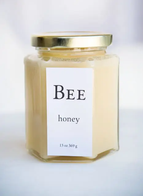 Hexagonal glass jar of light yellow honey with a gold lid and a white label reading "BEE honey 13 oz 369 g"