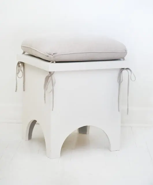 White storage bench with a light gray linen cushion tied with fabric ribbons on a white wooden floor.
