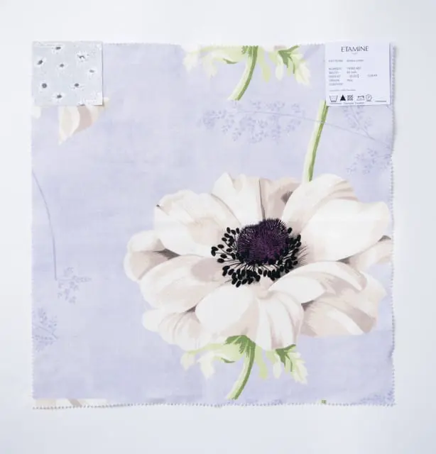 Fabric swatch with a large white anemone flower with a dark purple center on a light purple background. A small swatch of fabric with a grey floral pattern is attached to the upper left corner.