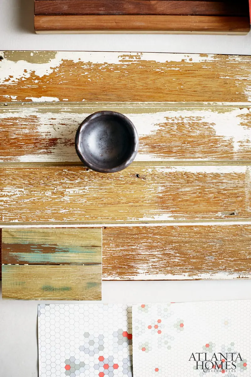 Close-up of a dark, rustic bowl on distressed wood planks with white paint peeling, next to wood samples and hexagon tile patterns, including an Atlanta Homes & Lifestyles logo.