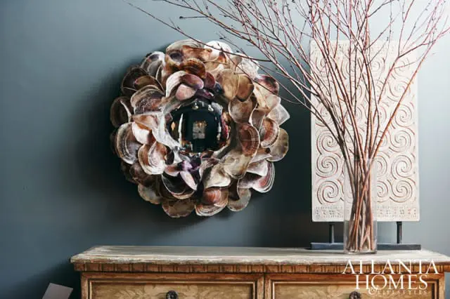 Round mirror with a frame of layered oyster shells in shades of brown and purple, displayed above a distressed wooden dresser with ornate drawer pulls.