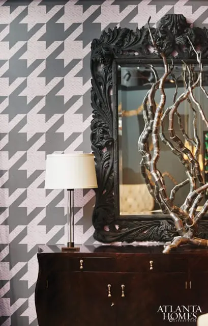 Dark wood credenza with brass hardware topped by a black ornate mirror and a silver branch sculpture. A table lamp stands to the left against a houndstooth wallpaper.