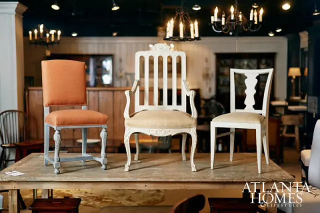 Three chairs displayed on a wooden table: an orange upholstered chair with carved gray legs, a cream upholstered armchair with ornate carvings, and a white upholstered chair with a decorative back.