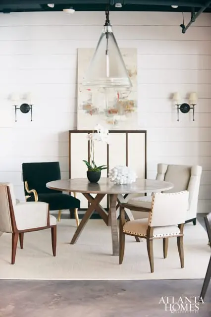 Dining room with a round wooden table, upholstered chairs in neutral tones and black velvet, a large glass pendant light, and abstract art above a cabinet.