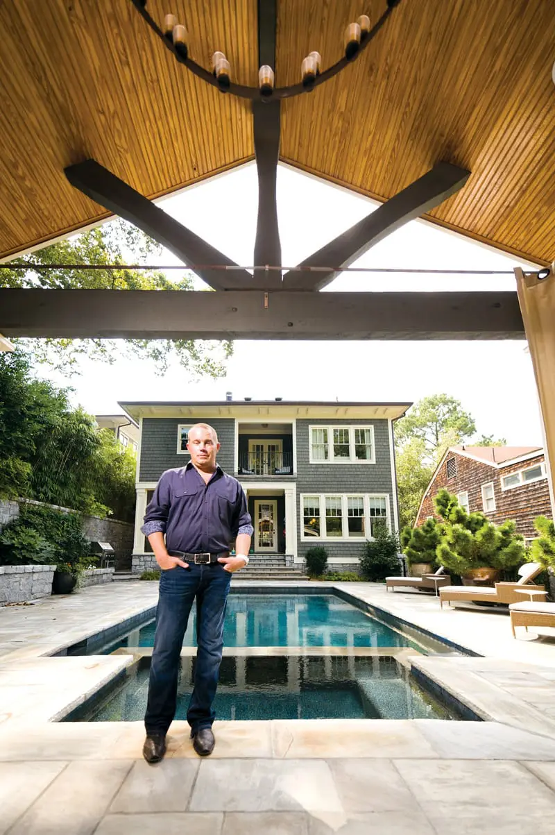 Man in a blue button-down shirt and jeans standing by a swimming pool in front of a gray house with white trim.