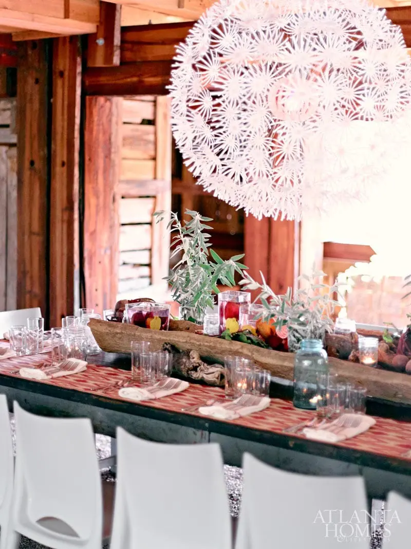 Rustic dining table set with white chairs, red patterned runner, glassware, candles, and a driftwood centerpiece with greenery and fruit.