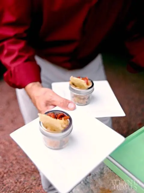 A person in a maroon shirt holds two white square plates, each topped with a small glass jar containing a bite-sized appetizer with a cracker and red topping.