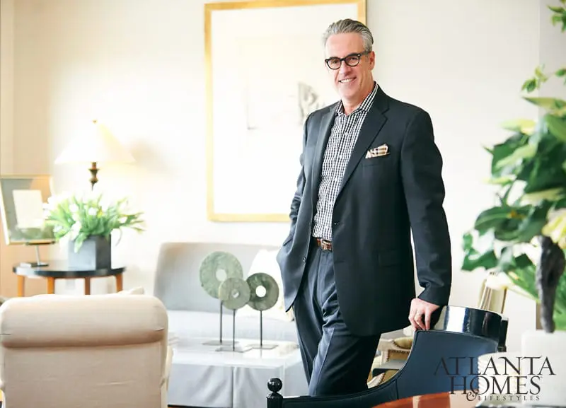 Man in a dark suit and checkered shirt stands in a living room with a gold-framed artwork behind him.