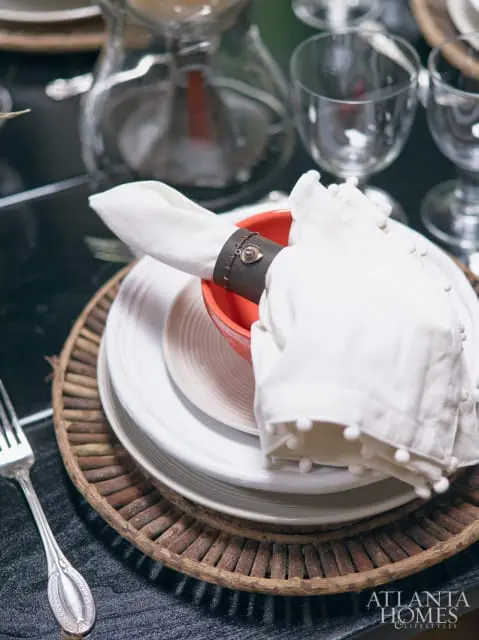 White napkin with pom-pom trim held by a leather napkin ring with a metal accent, set on white plates atop a rustic twig placemat.
