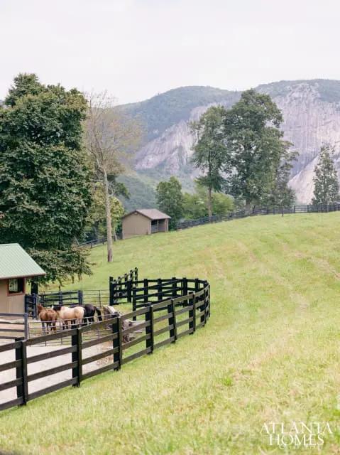 Pasture with horses in a black wooden fenced enclosure, a small barn, and a large mountain in the background under a cloudy sky.