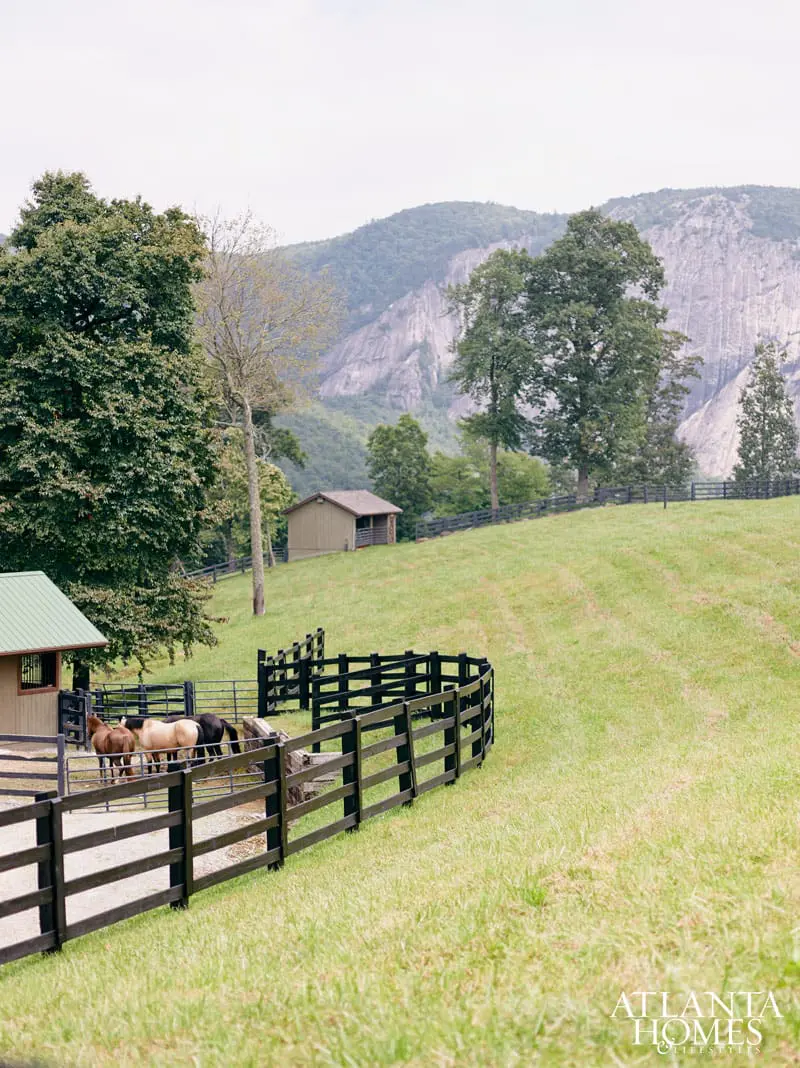 Pasture with horses in a black wooden fenced enclosure, a small barn, and a large mountain in the background under a cloudy sky.