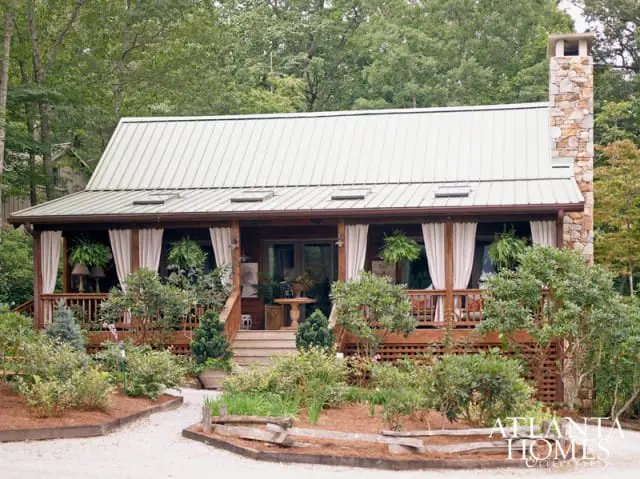 Log cabin with a metal roof, stone chimney, and covered porch with white curtains and hanging plants. Surrounded by lush greenery and landscaping.