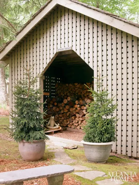Light beige wood shed with a slatted exterior, filled with stacked firewood. Two small evergreen trees in pots flank the entrance. A stone bench is in the foreground.