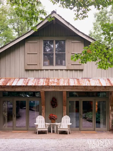 Rustic cabin exterior with weathered wood siding, corrugated metal roof, large window with shutters, and two white Adirondack chairs on a gravel patio.