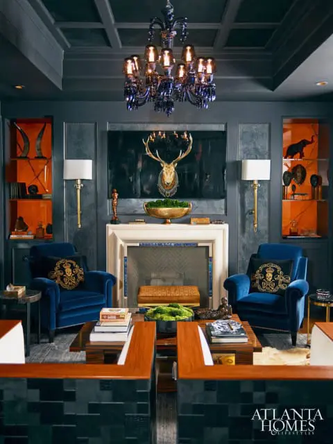 Dark gray living room with coffered ceiling, ornate chandelier, fireplace with mounted deer head, and two plush blue velvet armchairs with decorative pillows.