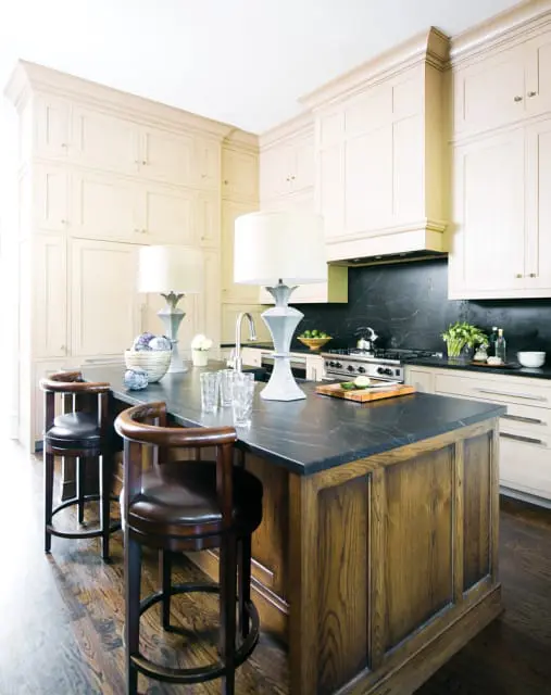 Cream kitchen with dark stone island topped with two lamps, dark wood paneled island base, and two brown leather bar stools.