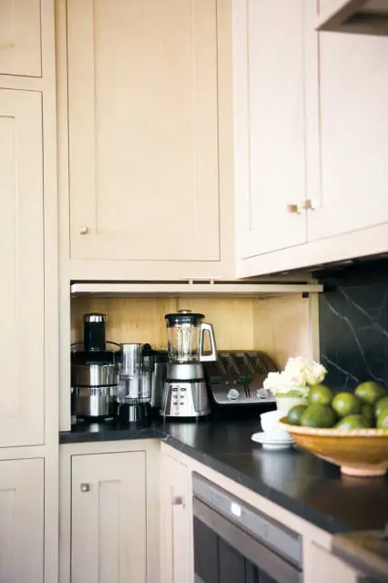 Cream kitchen with built-in appliance garage housing a blender, food processor, and toaster. Black countertop with a bowl of limes and white flowers.