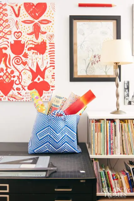 White wall with red abstract art and framed botanical print, blue woven basket filled with colorful rolled paper, and a bookshelf stocked with books.