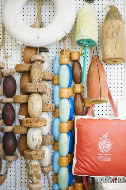 Collection of weathered wooden and cork buoys in various colors and sizes, hanging on a pegboard wall next to a life preserver and a red pillow with text.