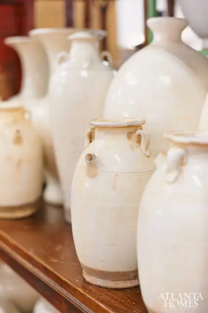Row of cream-colored ceramic vases with handles and spouts on a wooden shelf. Atlanta Homes & Lifestyles logo in the bottom right corner.