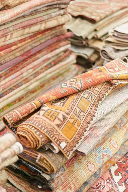 Stacks of rolled and folded oriental rugs in warm tones of orange, yellow, red, and beige with intricate geometric patterns.