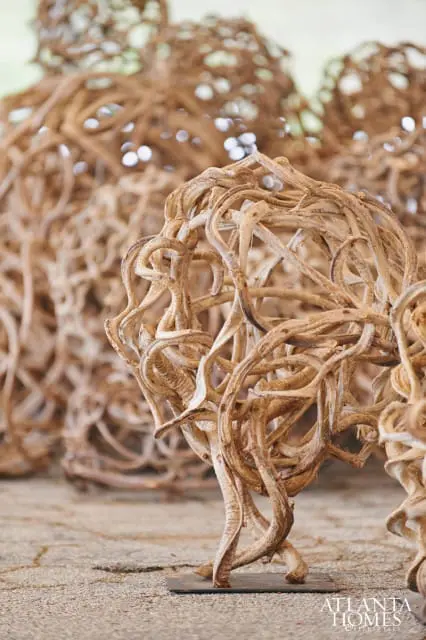 Close-up of a decorative sphere made of twisted, dried branches, resting on a dark metal base. Another similar sphere is blurred in the background.